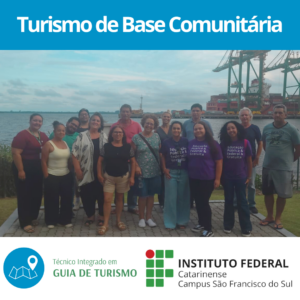 A imagem apresenta um grupo de pessoas reunidas ao ar livre em um espaço próximo a margem da Baía da Babitonga, com um porto e guindastes ao fundo. O céu está parcialmente nublado. O grupo é diverso, composto por homens e mulheres de diferentes idades e estilos de vestimenta. Algumas pessoas estão sorrindo, enquanto outras mantêm expressões mais neutras. Três mulheres no centro da foto vestem camisetas roxas com a frase "Educação Pública, Federal & Gratuita". O chão onde o grupo está posicionado é pavimentado com pedras e há uma faixa de grama entre eles e a água.
Na parte superior da imagem, há uma faixa azul com o texto "Turismo de Base Comunitária" em branco. Abaixo da foto, há uma barra branca com três elementos gráficos: um ícone azul de localização com um mapa, acompanhado do texto "Técnico Integrado em GUIA DE TURISMO" em verde; ao lado, o logotipo do Instituto Federal Catarinense Campus São Francisco do Sul.
