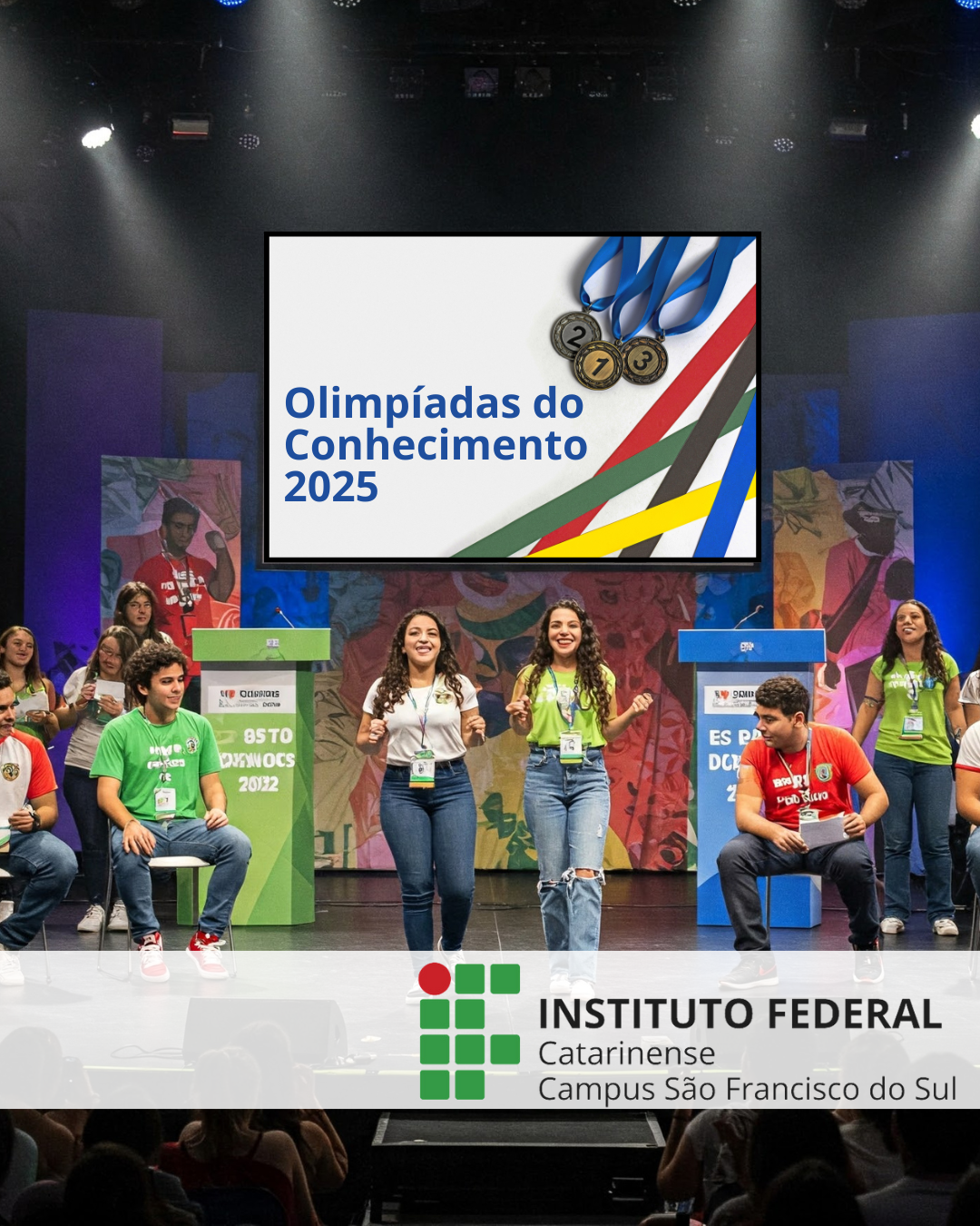 Em um palco iluminado por luzes de teatro, um grupo de jovens se apresenta diante de um público. Eles estão sorrindo e fazendo gestos animados. No fundo, um telão grande exibe o título "Olimpíadas do Conhecimento 2025", ao lado de três medalhas (ouro, prata e bronze) e faixas coloridas que lembram fitas olímpicas nas cores azul, vermelho, verde, amarelo e preto. No palco, há dois púlpitos — um verde e outro azul — cada um com um microfone e identificações de eventos passados. Jovens com camisetas de diferentes cores (verde, branco, vermelho) estão sentados ou de pé no palco, usando crachás pendurados no pescoço. Alguns usam camisetas com o nome do evento e logotipos educacionais. O fundo do palco é composto por painéis coloridos com grafismos artísticos e figuras humanas. Na parte inferior da imagem, há uma faixa translúcida com o logotipo do Instituto Federal Catarinense – Campus São Francisco do Sul.