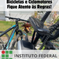 Imagem em formato vertical. Na parte superior, em destaque sobre uma faixa branca semitransparente, está escrito em letras grandes e pretas: "Uso e estacionamento de Bicicletas e Ciclomotores Fique Atento às Regras!" Ao fundo, aparece uma bicicleta preta estacionada junto a um ciclomotor, próximo a uma área gramada com vegetação e um muro claro. Na parte inferior da imagem está o logotipo do Instituto Federal Catarinense campus São Francisco do Sul.