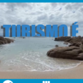Na parte superior da imagem, há uma faixa horizontal azul com o texto em branco: "10/05 Dia do Guia de Turismo". Abaixo da faixa, ocupa a maior parte da imagem uma fotografia de uma praia com mar calmo, céu nublado e pedras grandes nas laterais da areia. O mar avança suavemente até a faixa de areia em primeiro plano. No centro da fotografia, em destaque e em letras garrafais azuis, está escrito: "TURISMO É", com as letras parcialmente submersas e sombras projetadas na água, criando a sensação de profundidade. Na parte inferior da imagem, sobre outra faixa azul, há elementos institucionais. À esquerda, o ícone de um mapa com um marcador e os dizeres: "Técnico Integrado em GUIA DE TURISMO". À direita, o logotipo do Instituto Federal Catarinense campus São Francisco do Sul.