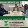 A imagem mostra três pessoas sentadas ao redor de uma mesa retangular clara, em uma sala de reuniões iluminada por luz natural que entra por uma janela ao fundo. À esquerda está o professor Adalto Aires Parada, diretor-geral do IFC São Francisco do Sul, vestindo uma jaqueta azul marinho e óculos. Ao centro está o Secretário de Turismo do município, Rangel Alexandre Friolin, de cabelos curtos, vestindo camiseta branca e casaco bege claro, com um caderno aberto à sua frente. À direita está a professora Patrícia Devantier Neuenfeldt, coordenadora da MICTI no campus, com cabelos castanhos claros e lisos, usando óculos e jaqueta jeans. Todos estão em conversa atenta. Na parte superior da imagem, há uma faixa verde com o texto em branco: “Parceria que fortalece a ciência e o turismo!” Na parte inferior, outra faixa verde contém o texto: “Representantes do Campus São Francisco do Sul participaram de uma reunião com o Secretário de Turismo do município, para articular o apoio da pasta à realização da XVIII MICTI.” Abaixo dessa faixa, aparece o logotipo do Instituto Federal Catarinense, com o texto: “INSTITUTO FEDERAL Catarinense Campus São Francisco do Sul” Ao fim da imagem, há o endereço do site do campus: saofranciscodosul.ifc.edu.br