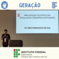 A imagem mostra o presidente do Grêmio Estudantil, ele está de camiseta bordô com crachá pendurado no pescoço, falando ao microfone em frente a um púlpito. Ao fundo, uma tela de apresentação exibe, em letras grandes, a frase: “8 – Implantação de Estrutura Modular de Convivência Estudantil – IFC São Francisco do Sul”. Na parte superior da imagem, há um banner com a logomarca do Grêmio Estudantil Geração. Na parte inferior, aparece o logotipo do Instituto Federal Catarinense – Campus São Francisco do Sul.
