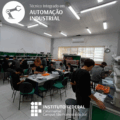 Laboratório de circuitos elétricos do campus São Francisco do Sul, com alunos distribuídos em mesas e bancadas de laboratório. Ao fundo, há estantes verdes com equipamentos eletroeletrônicos e luminárias articuladas. Os estudantes estão concentrados, manuseando peças e montando dispositivos. O logotipo do curso está na parte superior e o do IFC campus São Francisco do Sul no inferior.