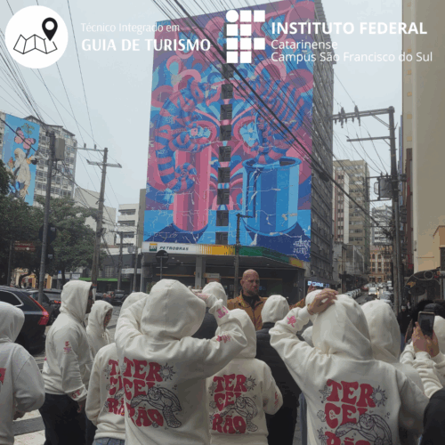 Fotografia de um grupo de estudantes, de costas, todos vestindo moletom branco com a palavra “TERCEIRÃO” em vermelho. Eles estão no centro de Florianópolis participando de um Street Art Tour, observando um grande mural colorido na lateral de um prédio. À esquerda, outro mural também aparece parcialmente. Na parte superior direita da imagem há o logotipo do Instituto Federal Catarinense – Campus São Francisco do Sul e, no canto superior esquerdo, um ícone de mapa com a inscrição “Técnico Integrado em Guia de Turismo”.