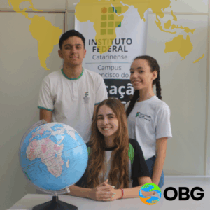 Três estudantes posam sorrindo em frente a um banner do Instituto Federal Catarinense – Campus São Francisco do Sul. Na mesa à frente deles há um globo terrestre azul. Uma estudante está sentada ao centro, e dois estudantes estão em pé atrás dela. No canto inferior direito está o logotipo colorido da OBG, e no topo da imagem há uma sobreposição gráfica em dourado com o mapa-múndi.