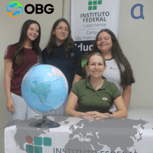 Quatro pessoas posam sorrindo em frente a um banner do Instituto Federal Catarinense – Campus São Francisco do Sul. Três estudantes estão em pé atrás e a professora Solange está sentada à frente, apoiando as mãos sobre a mesa, onde também há um globo terrestre azul. Na parte superior aparece o logotipo da Olimpíada Brasileira de Geografia – OBG, e da Associação Nacional de Pós-Graduação e Pesquisa em Geografia – ANPEGE, e da Olimpíada Brasileira de Geografia (OBG). Na parte inferior, aparece outra ilustração do mapa-múndi, desta vez em tom prata.