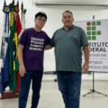 Dois homens posam lado a lado em um auditório do Instituto Federal Catarinense. Atrás deles há um conjunto de bandeiras e um banner institucional. Ambos sorriem em um ambiente claro e amplo.