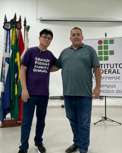 Dois homens posam lado a lado em um auditório do Instituto Federal Catarinense. Atrás deles há um conjunto de bandeiras e um banner institucional. Ambos sorriem em um ambiente claro e amplo.