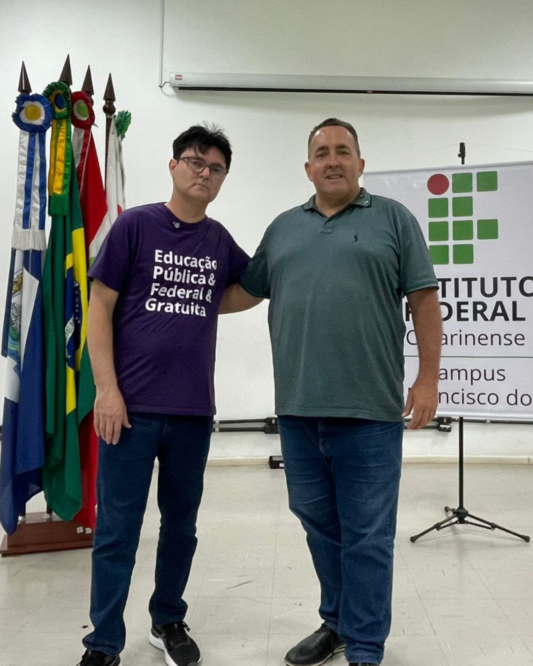 Dois homens posam lado a lado em um auditório do Instituto Federal Catarinense. Atrás deles há um conjunto de bandeiras e um banner institucional. Ambos sorriem em um ambiente claro e amplo.