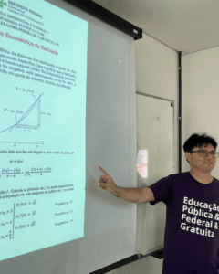Um homem de camiseta roxa apresenta uma aula projetada na parede. Ele aponta para o slide, que contém gráficos e fórmulas matemáticas, em uma sala de aula iluminada.